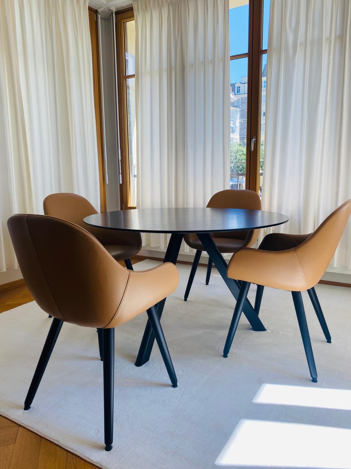 Séjour d’un appartement haussmannien à Lausanne : table ronde noire et quatre fauteuils en cuir cognac devant une rotonde vitrée